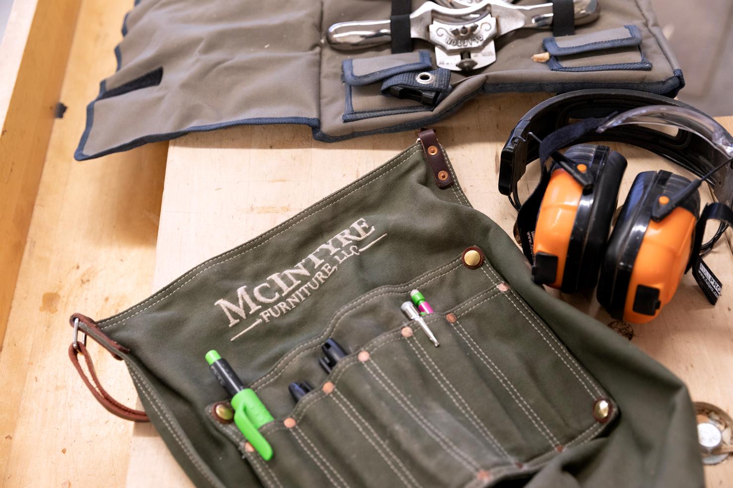 A work apron embroidered with "McIntyre Furniture, LLC" laying on a work bench next to ear protectors and other woodworking tools