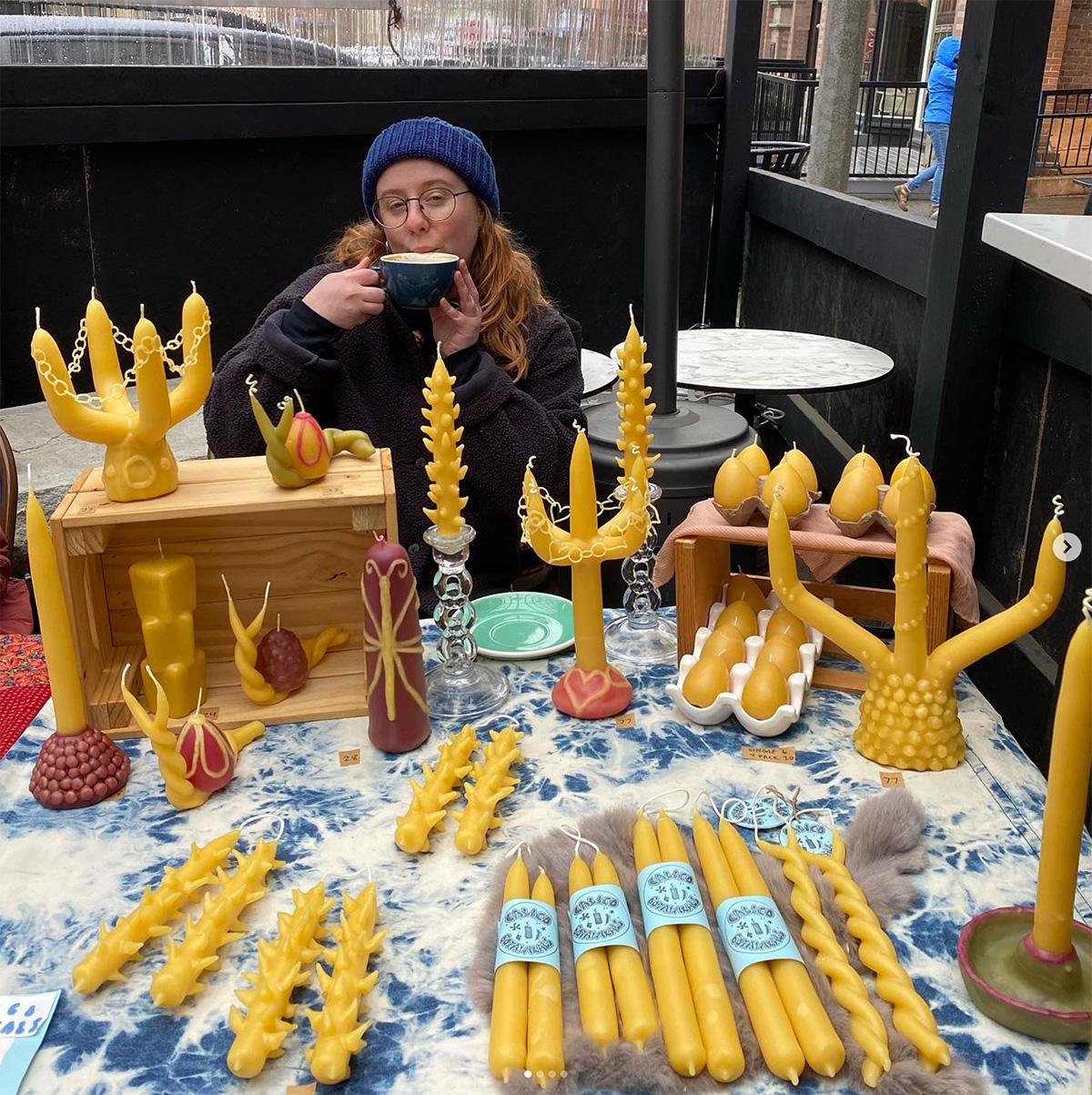 A woman sipping from a large mug behind a cable covered in eloborate candles