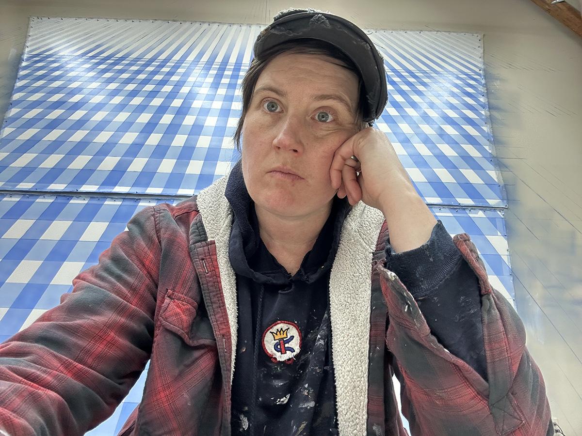 A person in a red flannel jacket and black hat resting their head on their hand with a blue and white artwork on the wall behind them