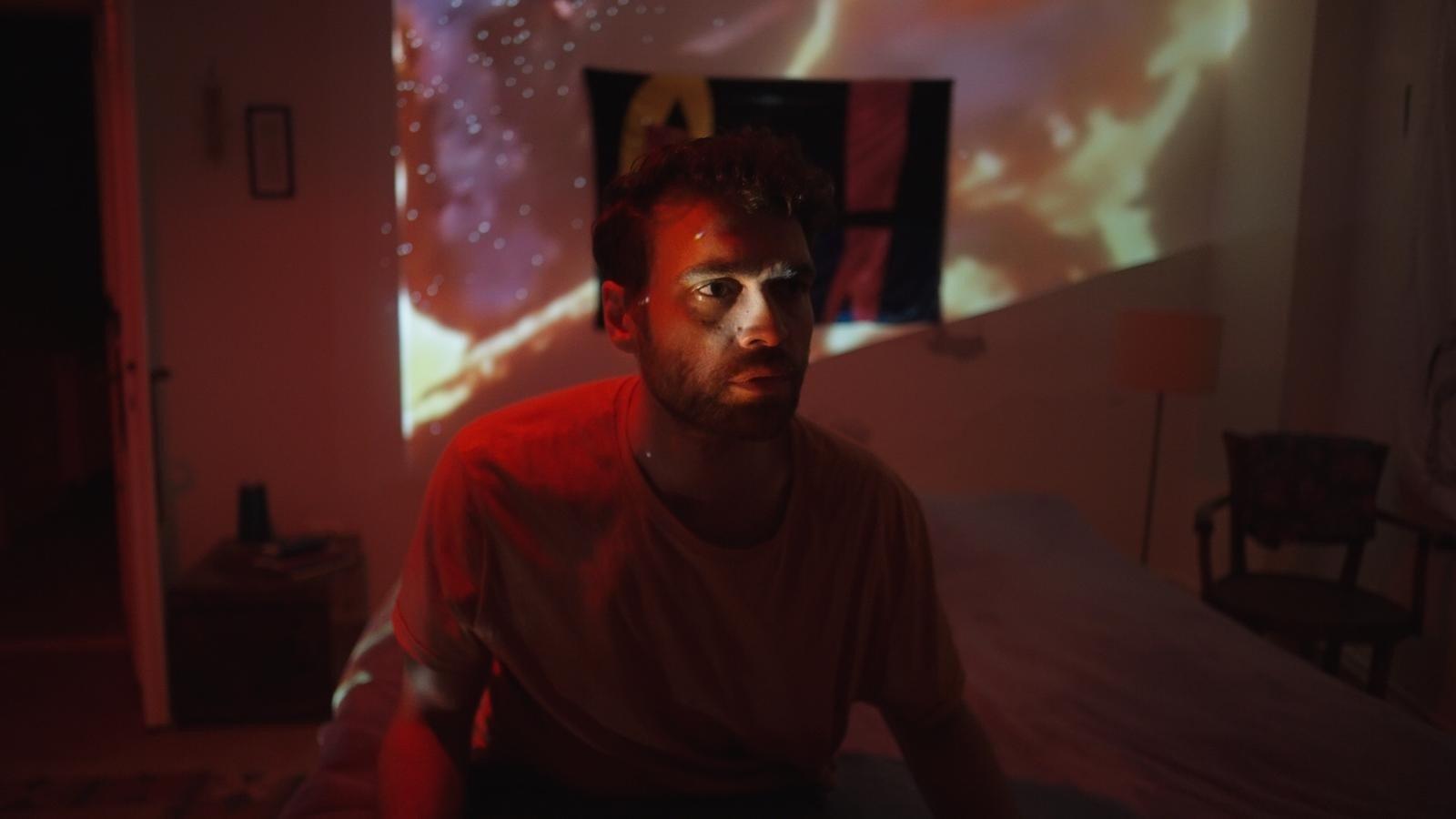 A man sitting on a bed in a bedroom lit up with red light and a projected image