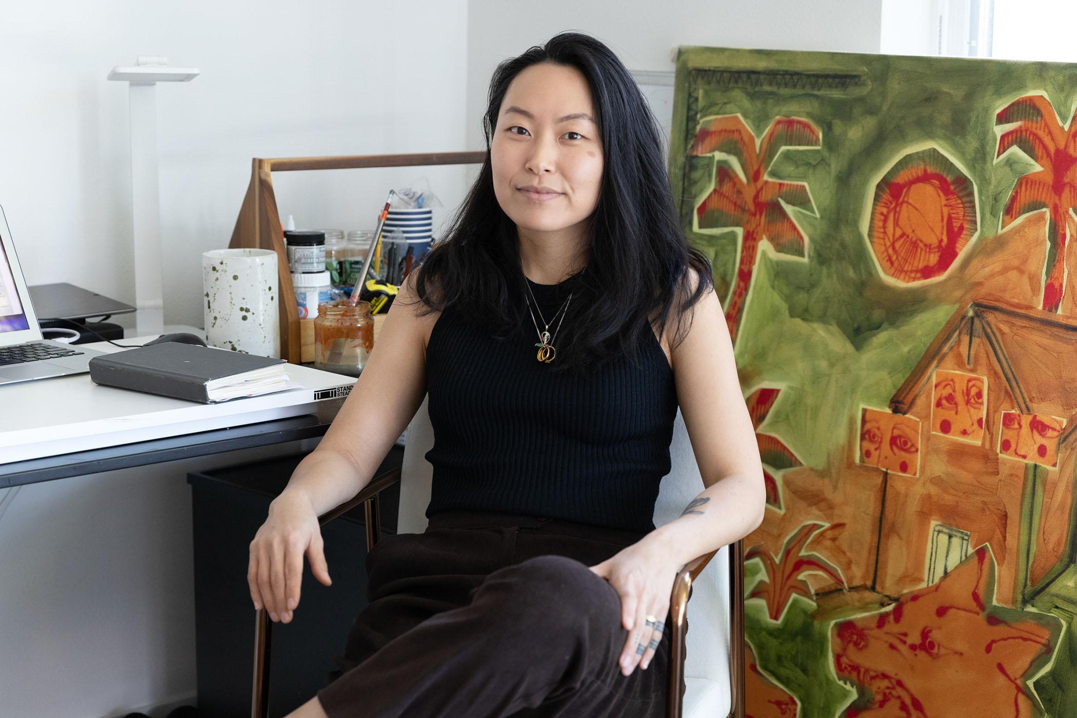An Asian woman wearing a black top and necklace sitting in front of a large artwork