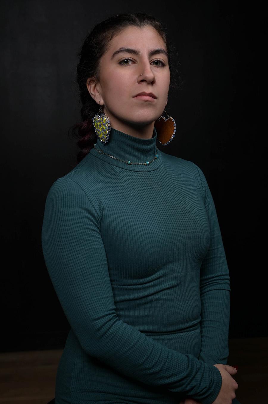 A professional headshot style photo of a person with large earrings and a longsleeve blue-green top against a dark background