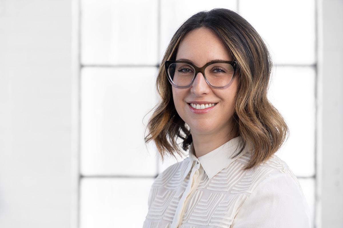 A professional headshot style photograph of a woman wearing glasses and a white blouse