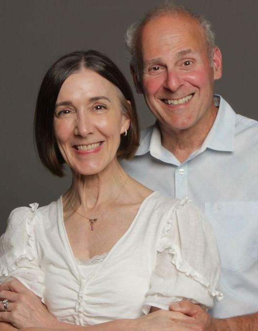 A professional headshot style photo of two people. In front is an older woman wearing a white shirt, and just behind and to the side is an older man wearing a light blue shirt.